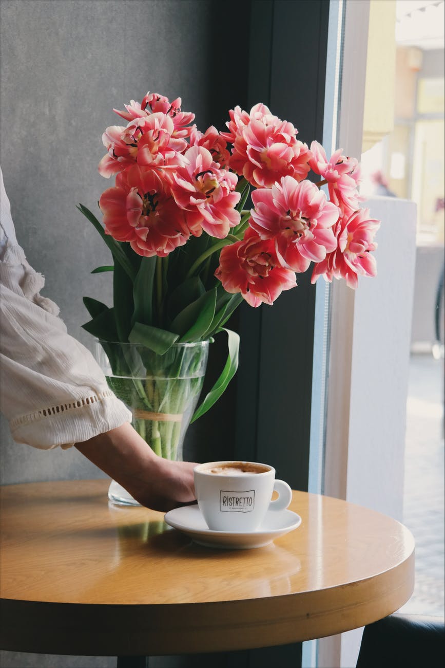 person placing cup of latte on white saucer near pink flowers in bloom